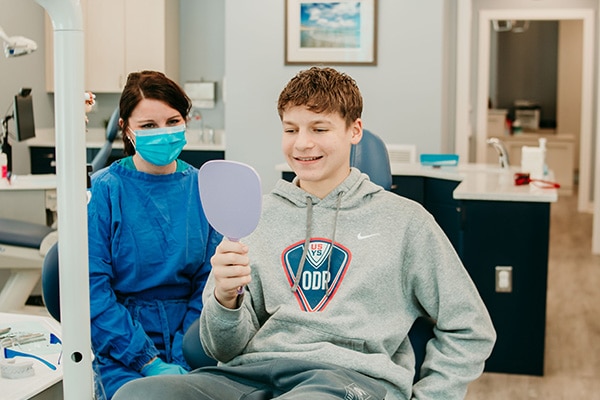 Teen patient smiling while looking into a handheld mirror during an Invisalign consultation, representing clear aligner treatment options at Griffin Orthodontics, Vernon, CT 06066.