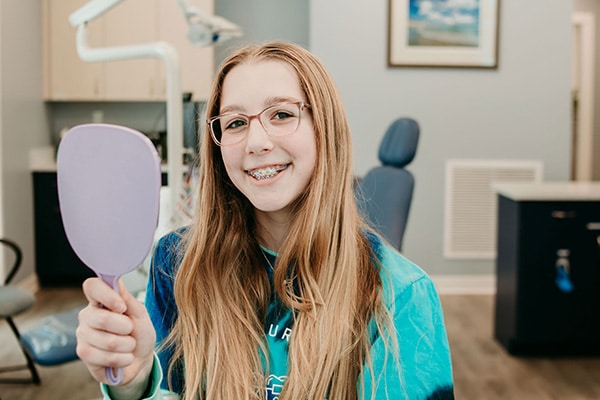 Smiling orthodontic patient with braces holding a handheld mirror in a treatment room, representing coordinated orthodontic care for complex bite concerns at Griffin Orthodontics, Vernon, CT 06066.